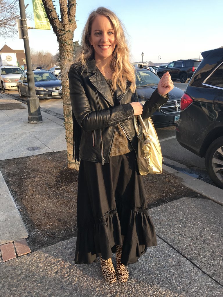 Woman in black skirt, leather jacket and gold purse.