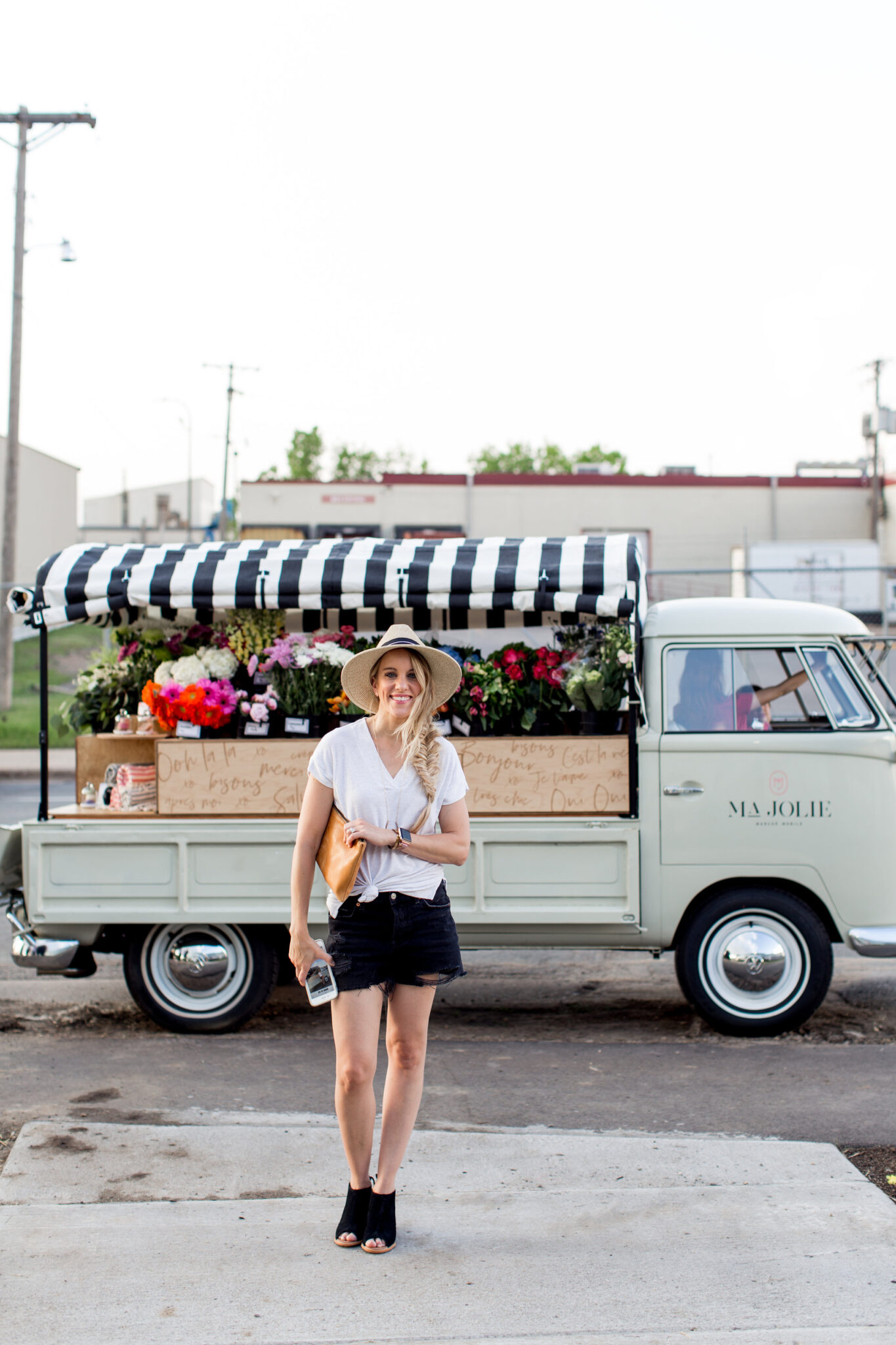 summer outfit for a casual day. blonde girl with fishtail braid and straw hat in front of majolie flower truck in black shorts, white top and black peep toe shoes