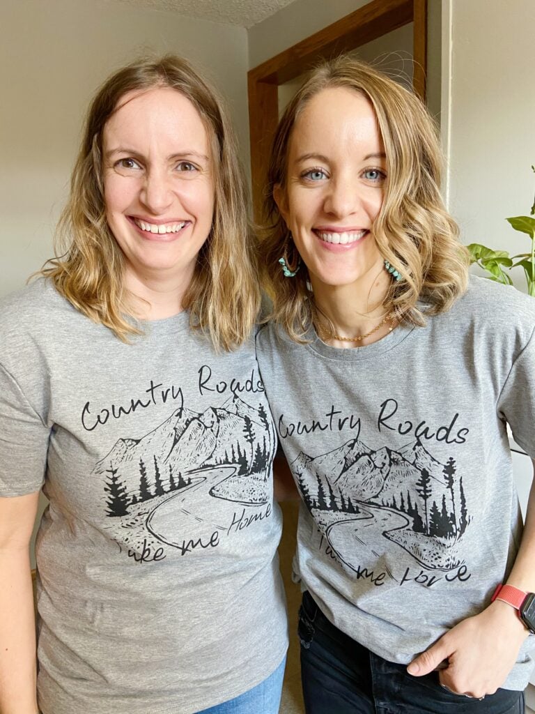 Two women wearing gray Country Roads Take Me Home tee