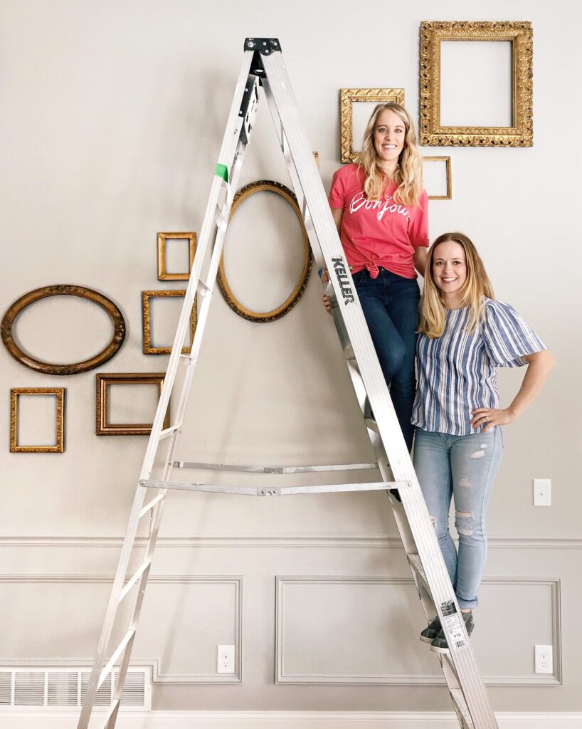 Two women on a ladder in front of a gallery wall of empty frames