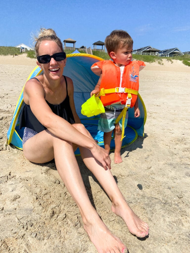 mom and baby in life jacket on the beach next to small beach tent