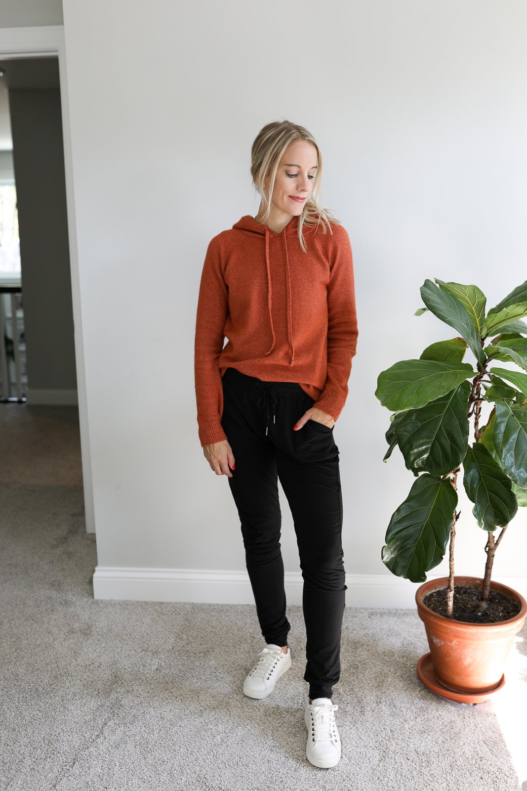 woman wearing white sneakers, black joggers and orange hooded sweater standing next to fiddle leaf fig tree