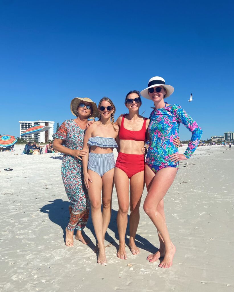 four women at the beach - one in a caftan, two in strapless high waisted bikinis and one in a rash guard suit with a hat