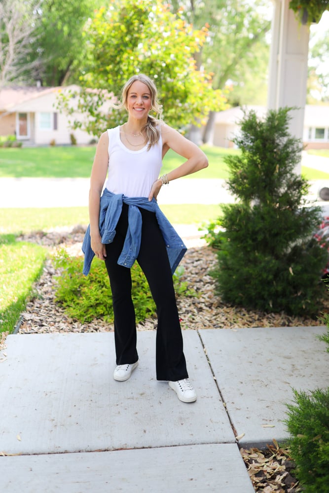 Woman wearing flare leggings with white tank with chambray shirt around waist and white sneakers.