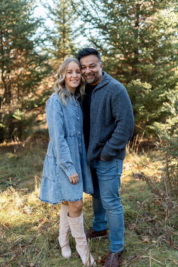 woman in denim dress and man in blue cardigan outside in front of trees