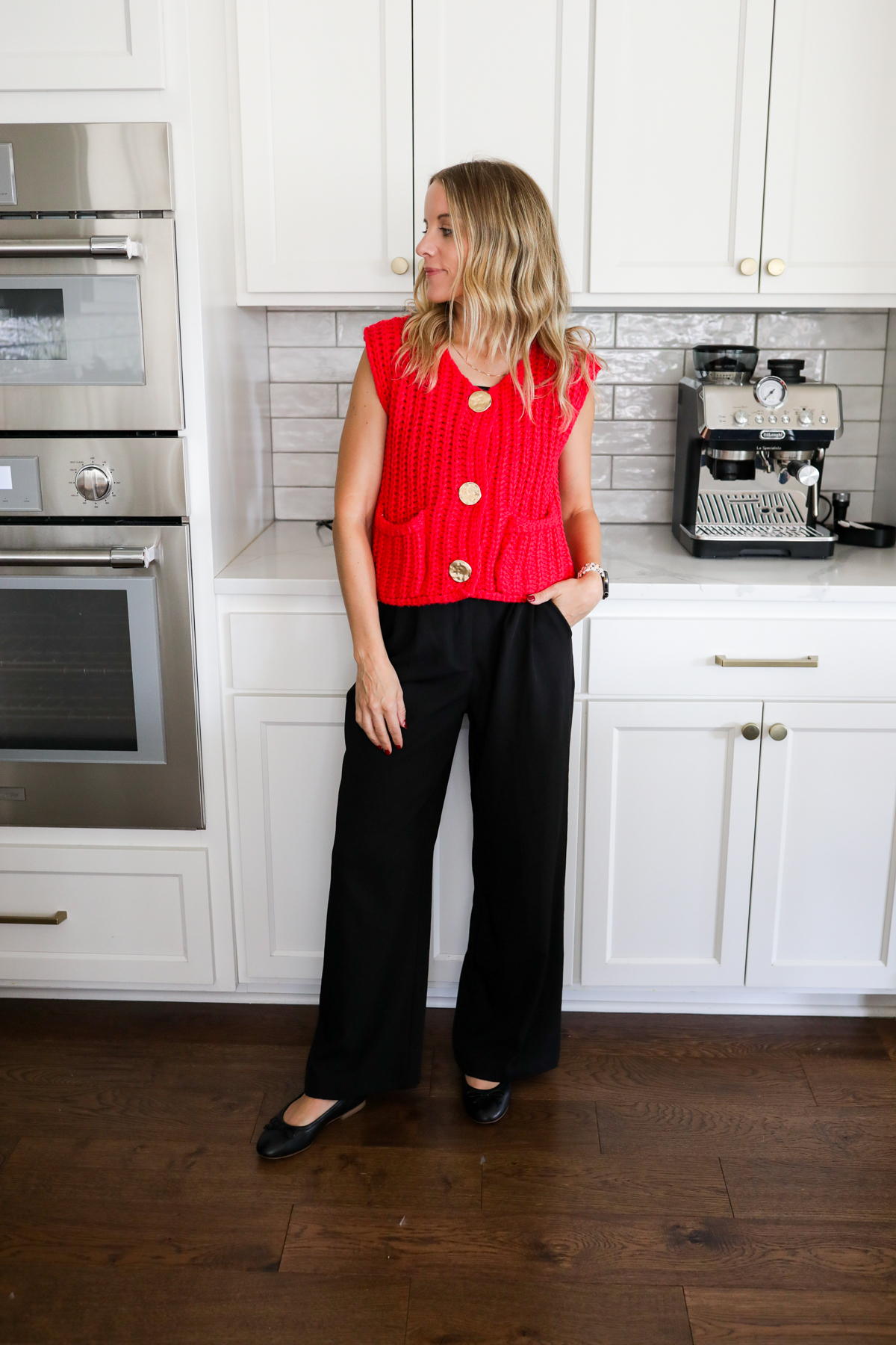 Woman in black trousers and a red sweater tank standing in a kitchen.