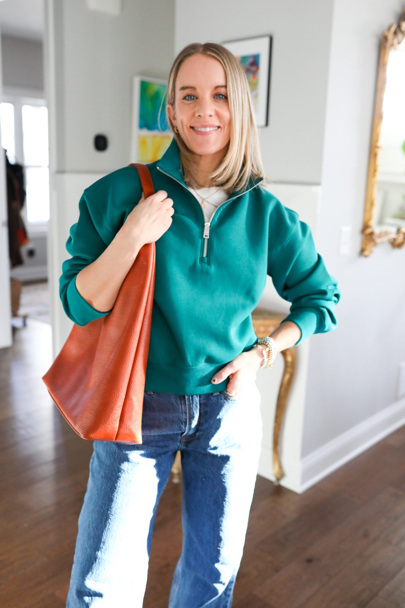 woman wearing green half zip pullover, dark wash jeans, and brown tote bag.