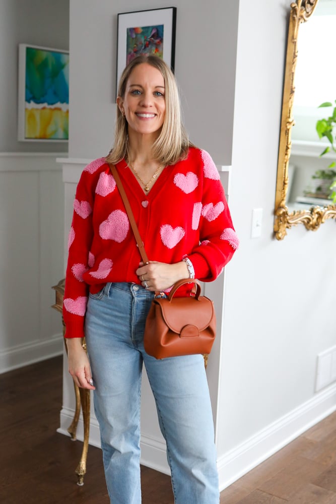 Woman wearing red valentine's day sweater cardigan, brown cross body bag, and jeans. 
