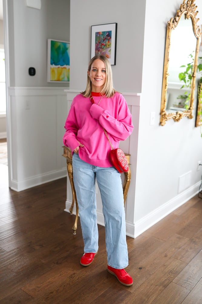 Woman wearing pink valentine's day sweatshirt, jeans, red shoes, and red cross body bag.