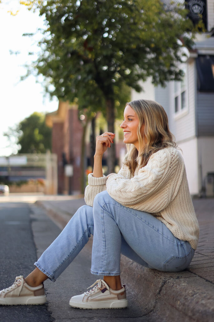 Woman in jeans and a sweater sitting on the curb.