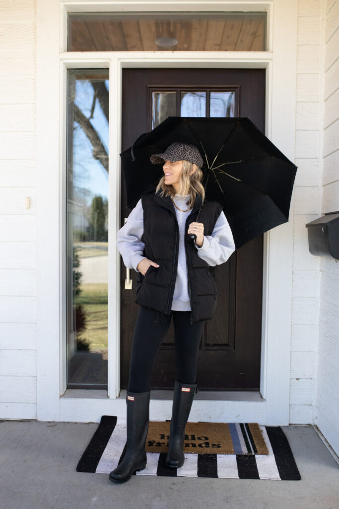 Woman in back leggings with a grey sweatshirt and black vest paired with Hunter boots holding an umbrella