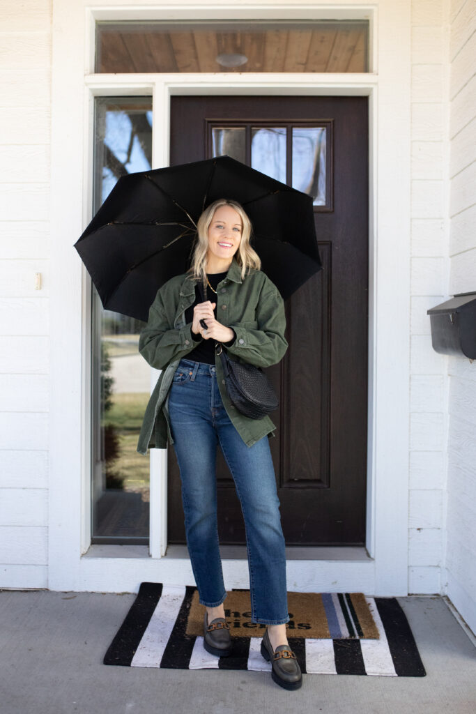 Woman in jeans with a black top and green jacket paired with loafers holding an umbrella