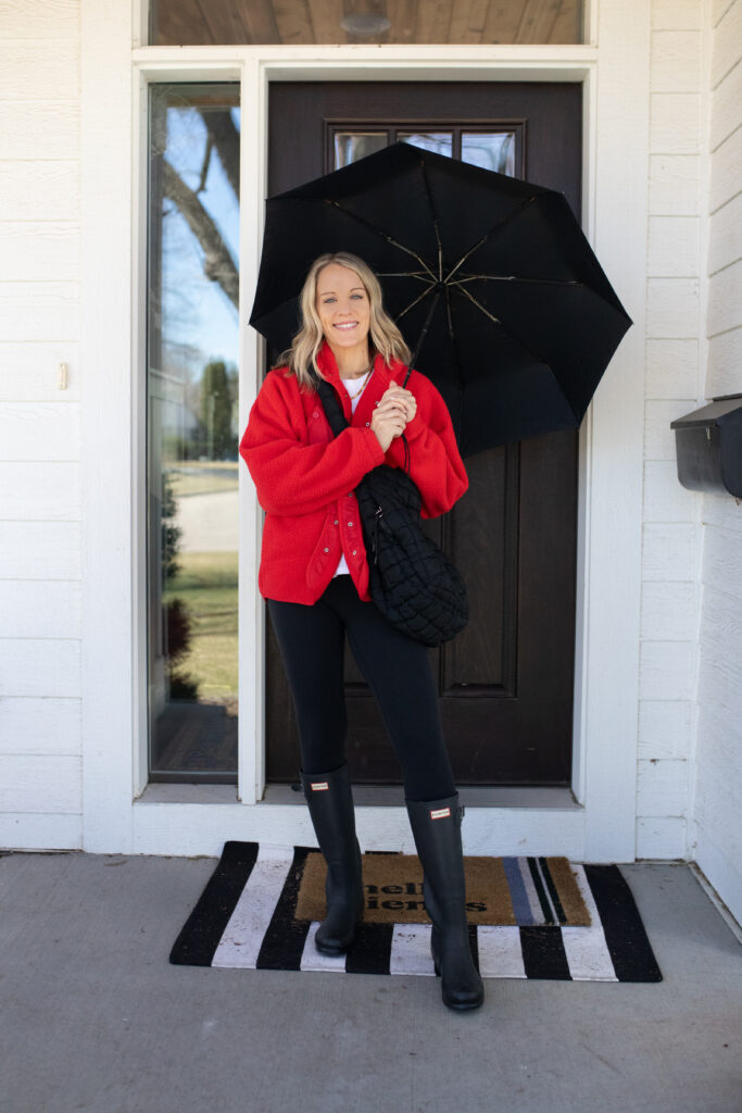 Woman with a white tee and red jacket and black leggings paired with Hunter brand rain boots and an umbrella