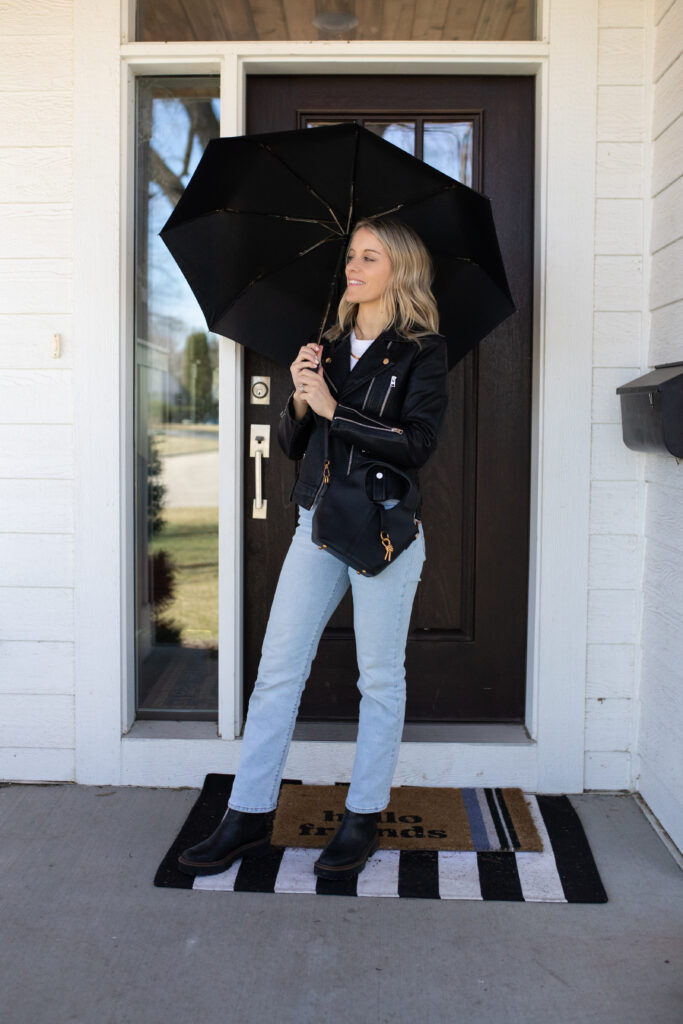 Woman in light wash jeans with a white tee and black jacket paired with dark boots and black bag holding an umbrella