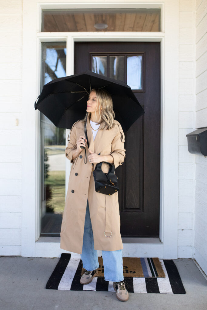 Woman in light wash jeans with a white tee and beige rain jacket paired with tennis shoes holding an umbrella