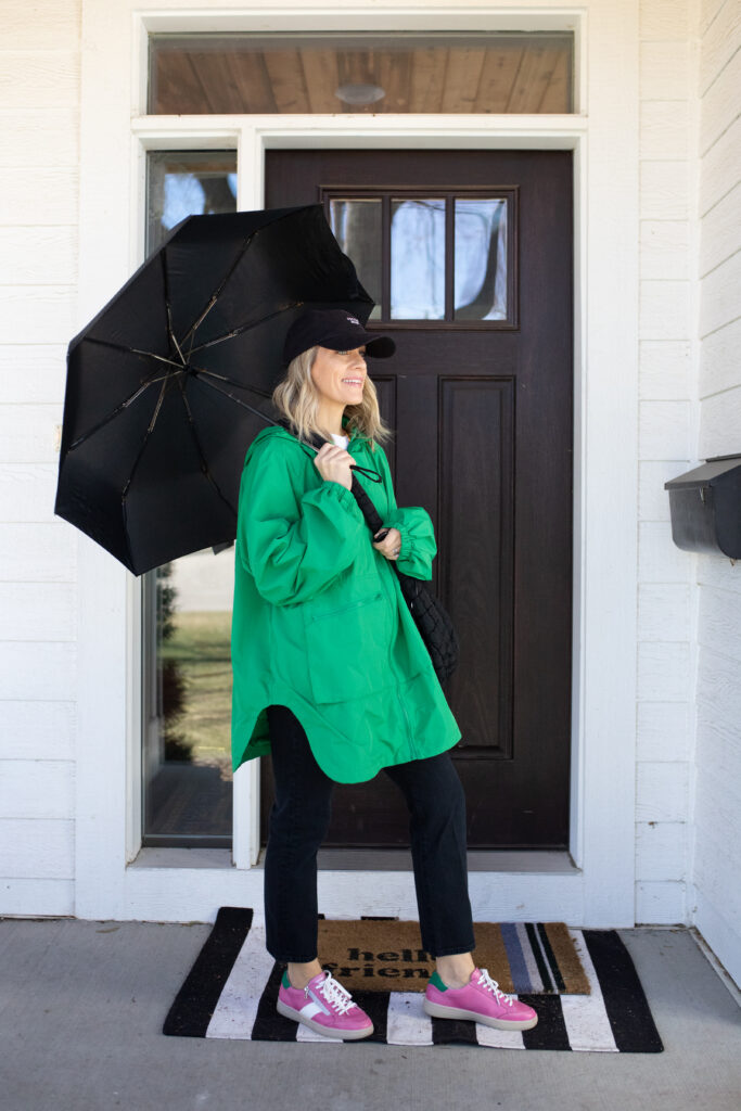 Woman in green rain jacket and black pants with pink sneakers and a black baseball cap holding a black umbrella