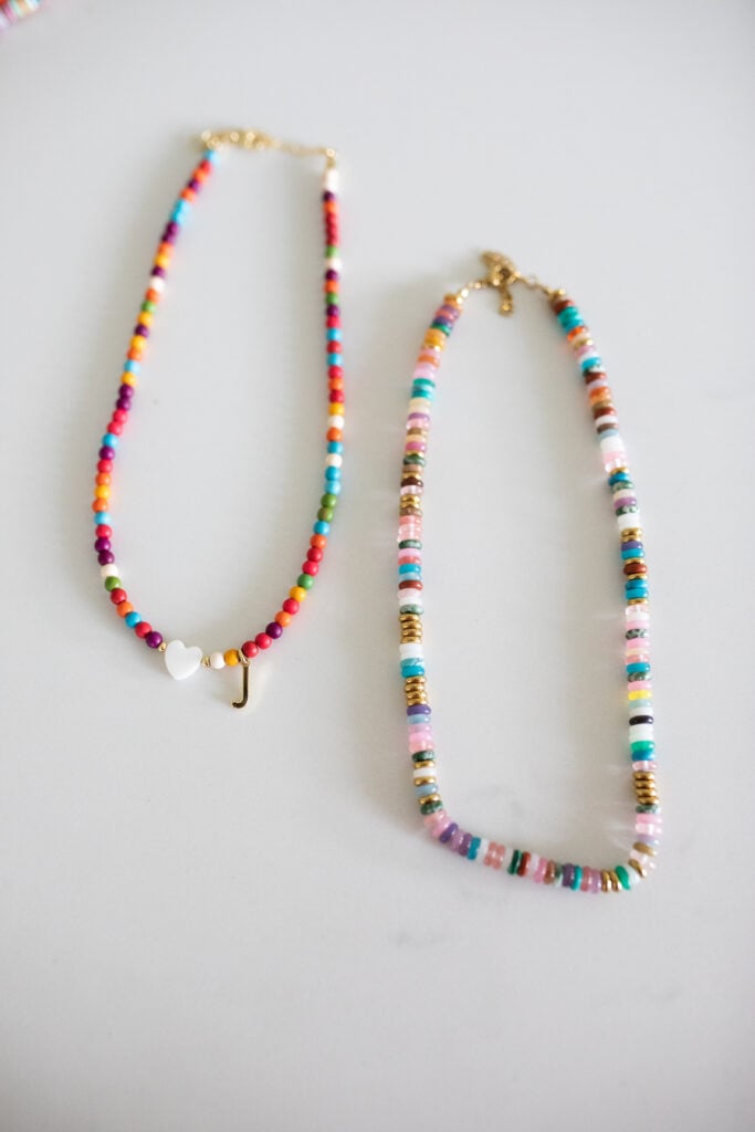 A close-up of two trendy beaded necklaces ā one with a customizable letter pendant and the other with vibrant beads all around, adding fun color and personality.