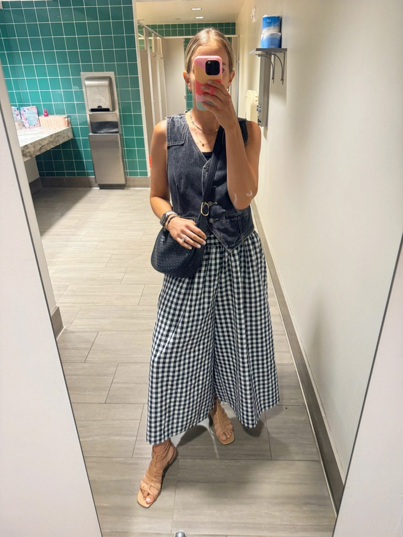 n Paisley of Paisley and Sparrow in a chic street style outfit featuring a black washed denim vest, layered silver and gold necklaces, black woven crossbody bag, and gingham wide-leg pants.