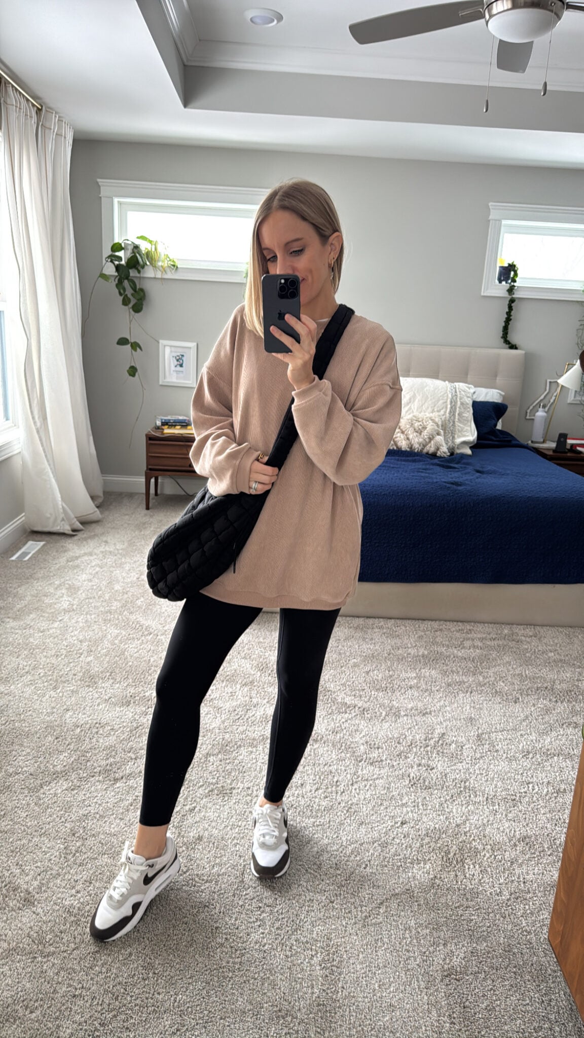 Woman taking a mirror selfie in a neutral Shewin oversized sweatshirt paired with black Aoxjox leggings. Styled with white and gray Nike sneakers and a quilted black crossbody bag for a cozy, casual look.