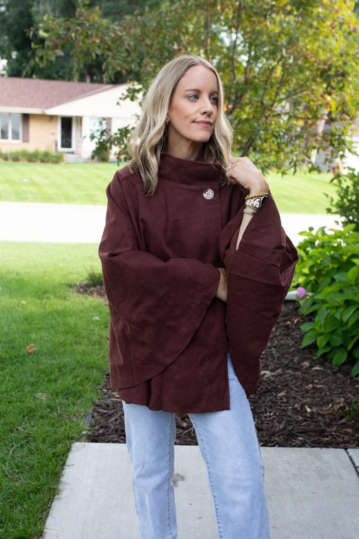 Jen Paisley of Paisley and Sparrow wearing a burgundy cape coat with wide sleeves styled for fall fashion, paired with light wash denim jeans, outdoor street style look.
