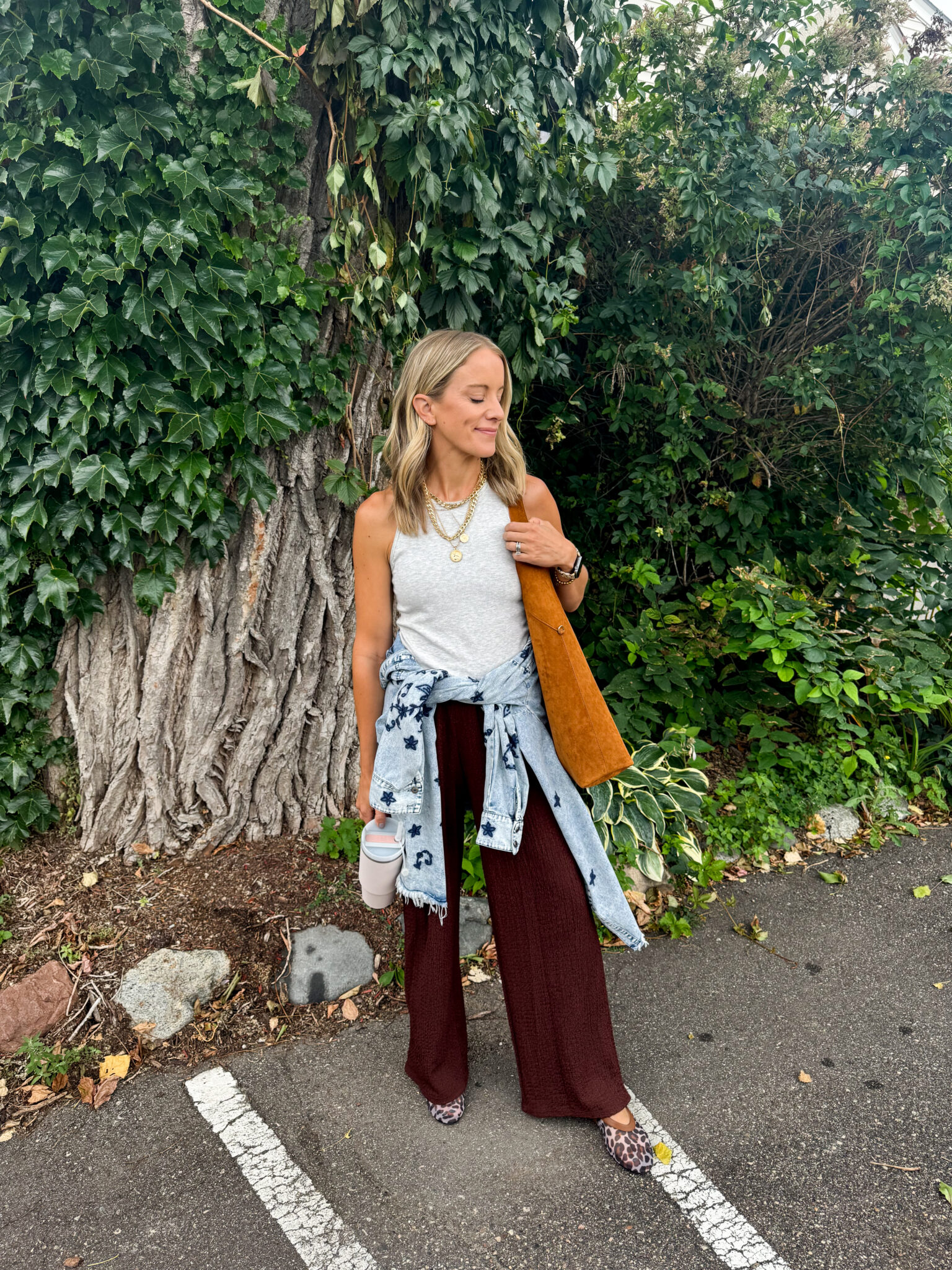 Jen Paisley of Paisley and Sparrow in a casual fall outfit with chocolate brown wide-leg pants from Target, light grey Vuori tank top, suede brown tote bag, leopard flats, and a denim jacket tied at the waist.