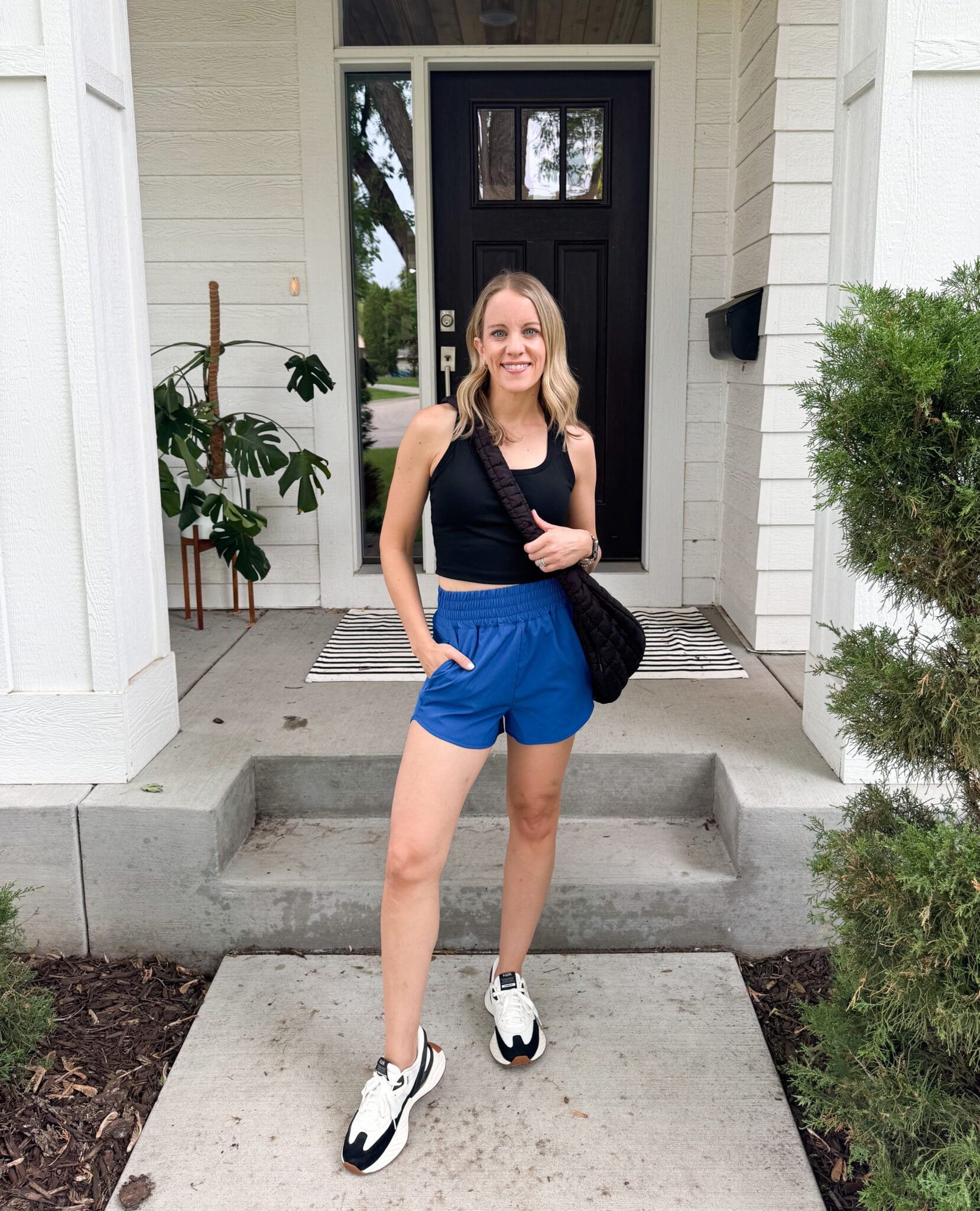Jen standing on a front porch wearing a black athletic crop tank and cobalt blue Pinspark shorts. Paired with white and black sneakers and a crossbody bag .