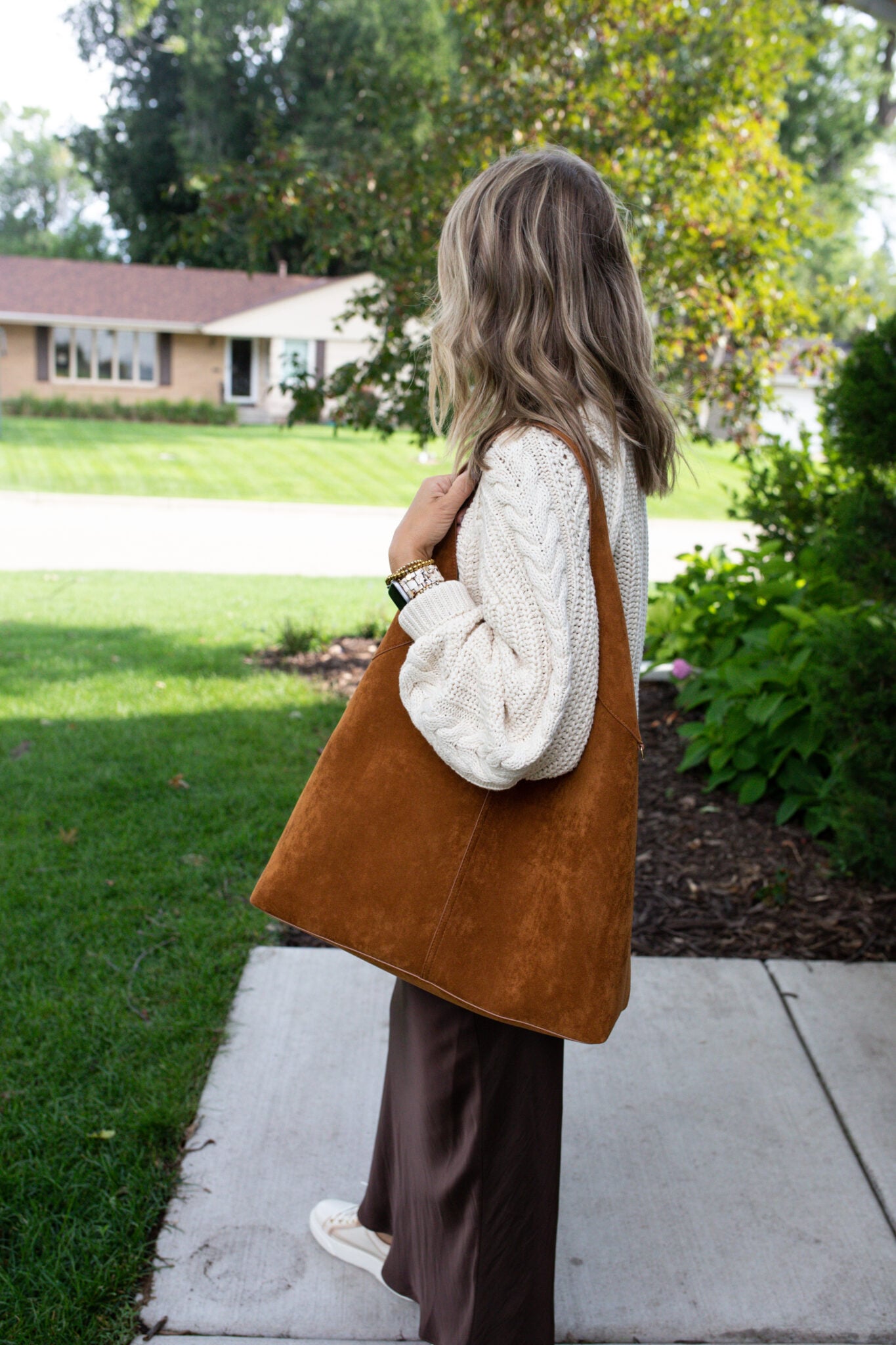 Jen Paisley of Paisley and Sparrow showing a side view of a fall fashion look featuring a large tan suede slouch bag from Amazon, paired with a cream knit sweater and chocolate brown midi skirt.
