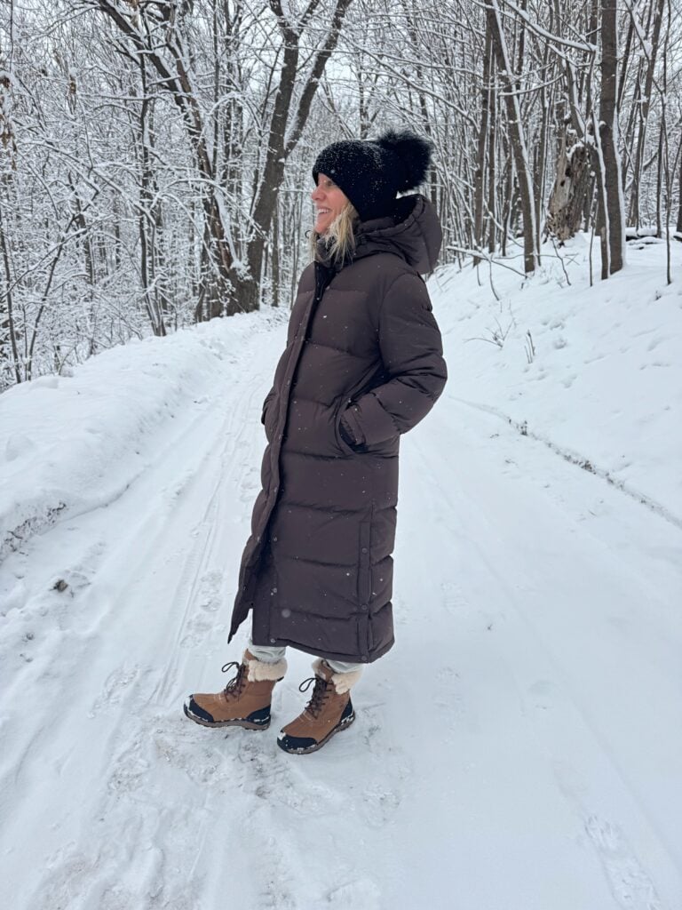 Woman outside in the snow wearing a hat, long puffer jacket, and boots.