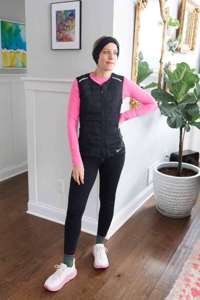 Woman in pink running shirt, black vest, black leggings and sneakers.